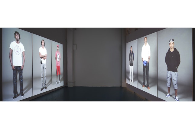 Installation view of Stop, a two-channel projected HD video installation. A video projection on one wall features young men from Brooklyn, New York, while the video on the facing wall features young men from Liverpool, England. Each young man repeatedly states the number of times he has been stopped by the police. Photo credit: © Dread Scott Room with multiple projections on wall