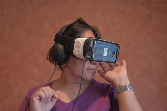 From the series “Fractured Connections” A Family Reunions Project participant explores the neighborhood where she once lived in Puebla, Mexico. Paterson, New Jersey, United States, 2017. Photo credit: © FRPxTN A woman wearing a virtual reality headset