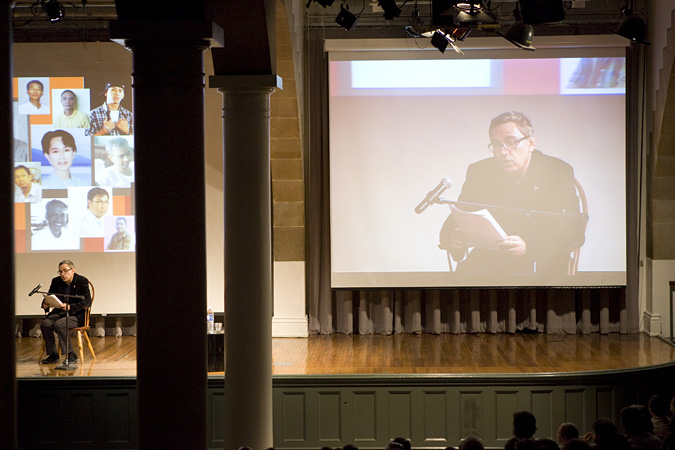 Orhan Pamuk reading into microphone; large screen in the background