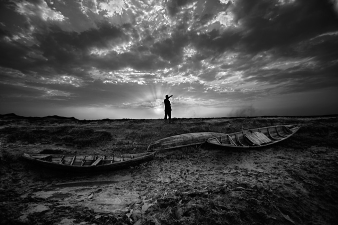 Man standing on banks points beyond some boats on land