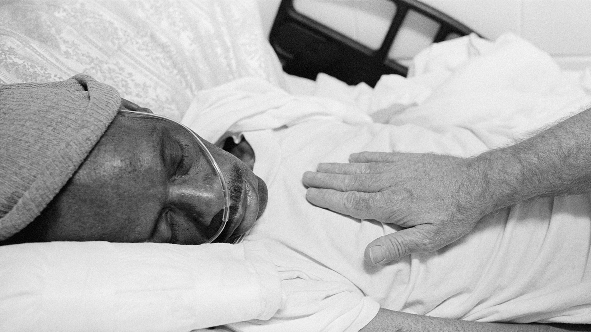 A man lays in a hospital bed with someone's hand on his chest