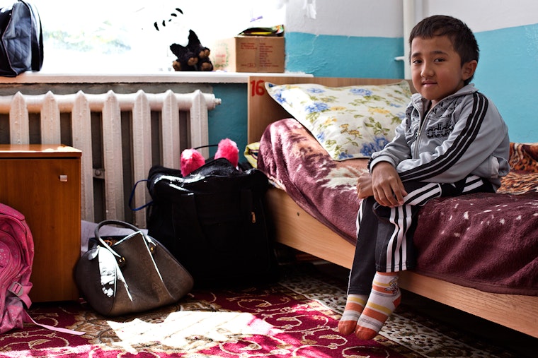 Askar sits in his room at an orphanage in Kyrgyzstan. He says, “When I came to class, I was fascinated by my teacher, books, a desk, and all things you need to study. It was very difficult to learn to write in the beginning, but then it got better.” Photo credit: © Ellie Kealey A young boy on his bed