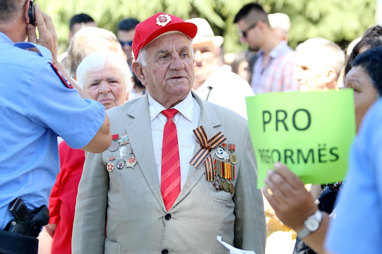 An elderly man with military insignia on his jacket stands amidst a crowd during a sunny day.