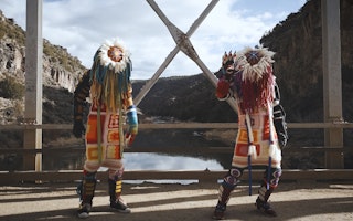Two figures stand on a bridge dressed in hand-made Indigenous regalia.