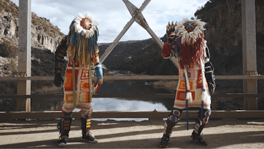 Two figures stand on a bridge dressed in hand-made Indigenous regalia.