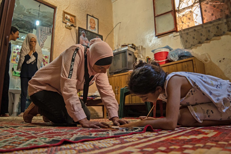 A refugee organizer visits a refugee family to check on the children’s school work in the Shatila refugee camp in Lebanon on August 23, 2020. Photo credit: © Ali AlSheikh Khedr for the Institute of International Education A person visits with a young girl, who is writing with a pencil.