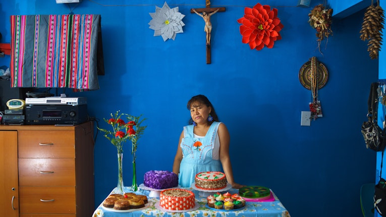 From the series “Project Luz” Roberta Chalini is a member of Mujeres en Movimiento, a self-organized group that explores the use of dance, art, ancestral remedies, and civic engagement to empower Latina immigrant women in Corona, Queens. This portrait of Roberta and her cakes was created during a collaborative portraiture workshop. It is a representation of her dream of making cakes for other people coming true. Queens, New York, United States, 2018.	Photo credit: © Project Luz A woman standing in front of a deep blue wall