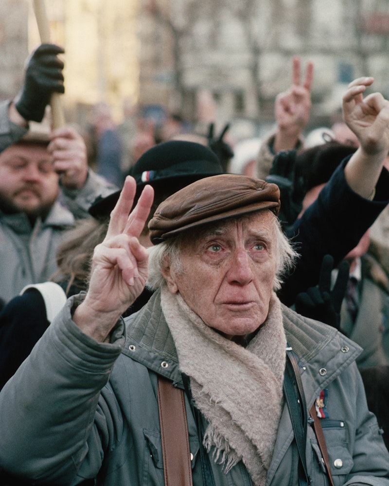 An older man standing in a crowd holding two fingers in the air