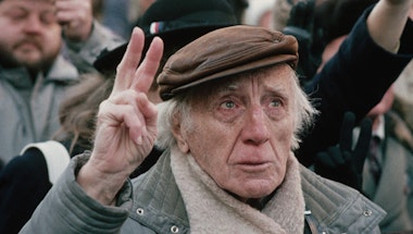An older man standing in a crowd holding two fingers in the air