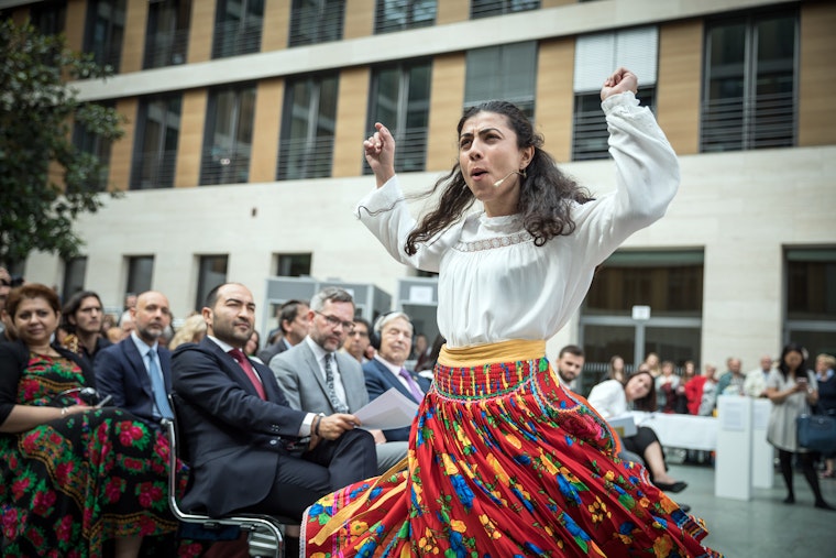 An actor performs at the launch of the European Roma Institute for Arts and Culture in Berlin, Germany. Photo credit: © Gordon Welters/Laif/Redux for the Open Society Foundations A woman is spinning in a floral skirt among an audience of people in large atrium hall.