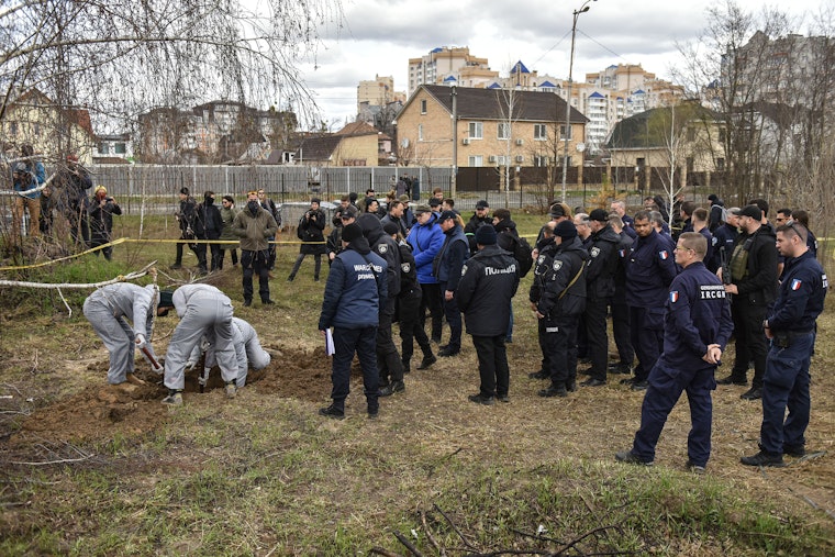 Members of an international team of war crimes prosecutors visit a mass grave in Bucha, Ukraine, on April 12, 2022. Photo credit: © Oleg Petrasyuk/EPA-EFE/Shutterstock A group of people gathered around people digging in a neighborhood lot