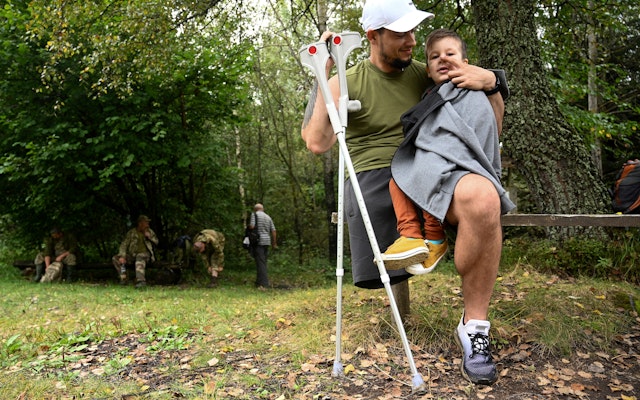 A veteran and his son rest after a hike assisted by volunteers in Trukhaniv, Ukraine, on September 16, 2023. © Yuriy Dyachyshyn/AFP/Getty A man with crutches sits with his son on his lap