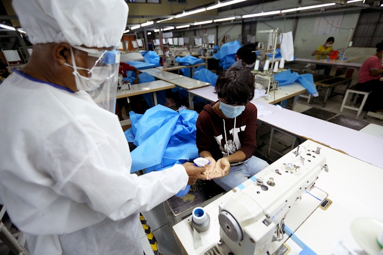 A person in full protective gear sprays the hands of a worker sitting at a sewing machine