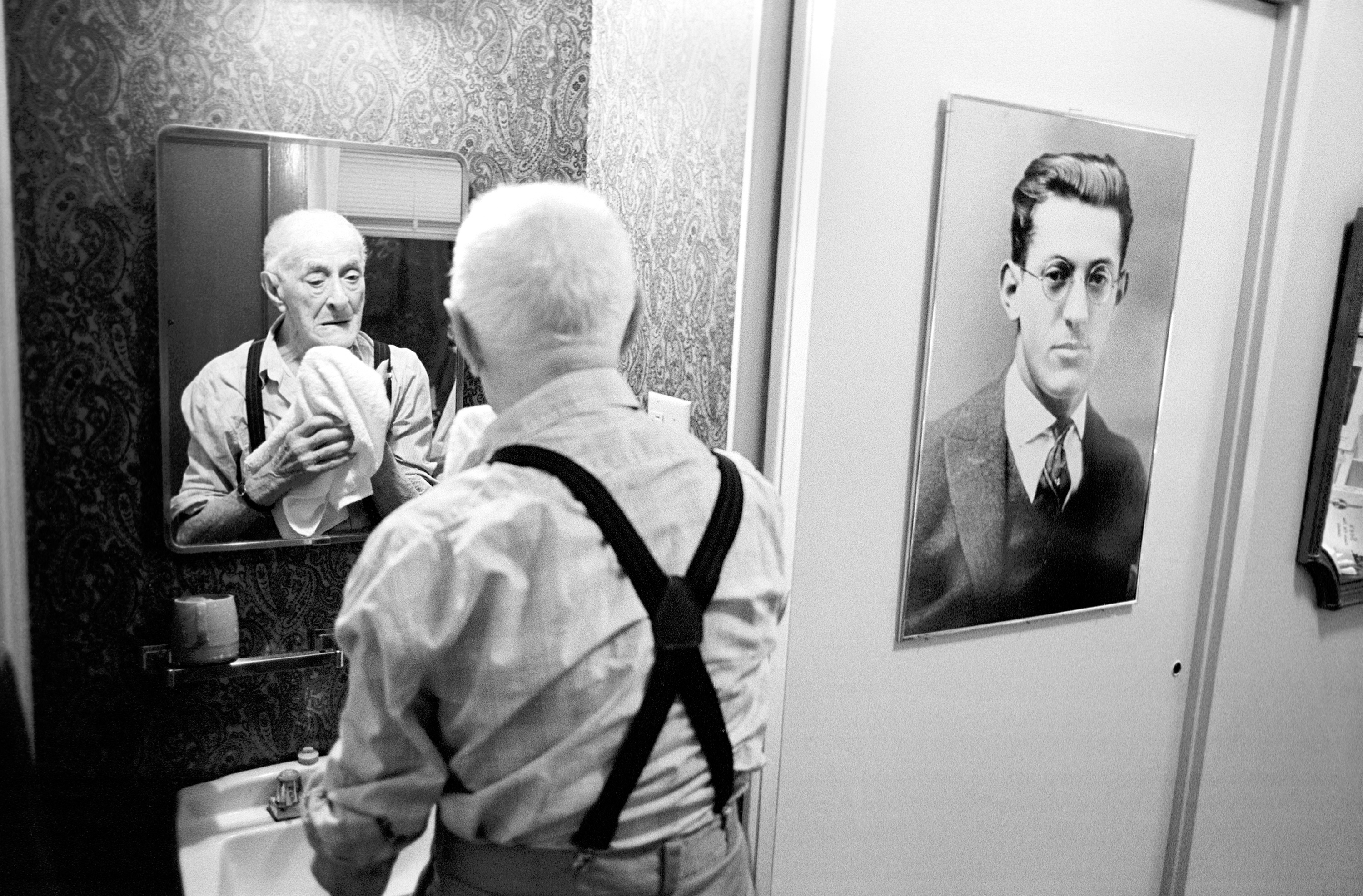 An older man washes his face in the mirror next to a portrait of his younger self