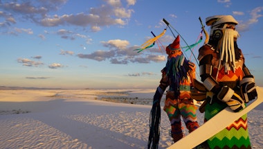Two figures stand in front of a landscape dressed in hand-made Indigenous regalia.