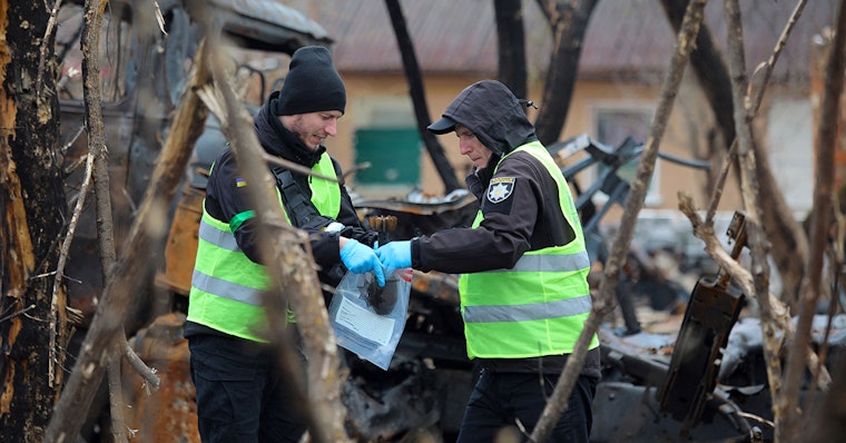 Ukrainian police forensic investigators examine an area with burnt Russian military vehicles in Bervytsia, Ukraine, on April 21, 2022. Photo credit: © Aleksey Filippov/AFP/Getty Two people in neon vests place an object in a bag