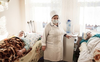 A nurse stands between two patients in beds in a hospital room