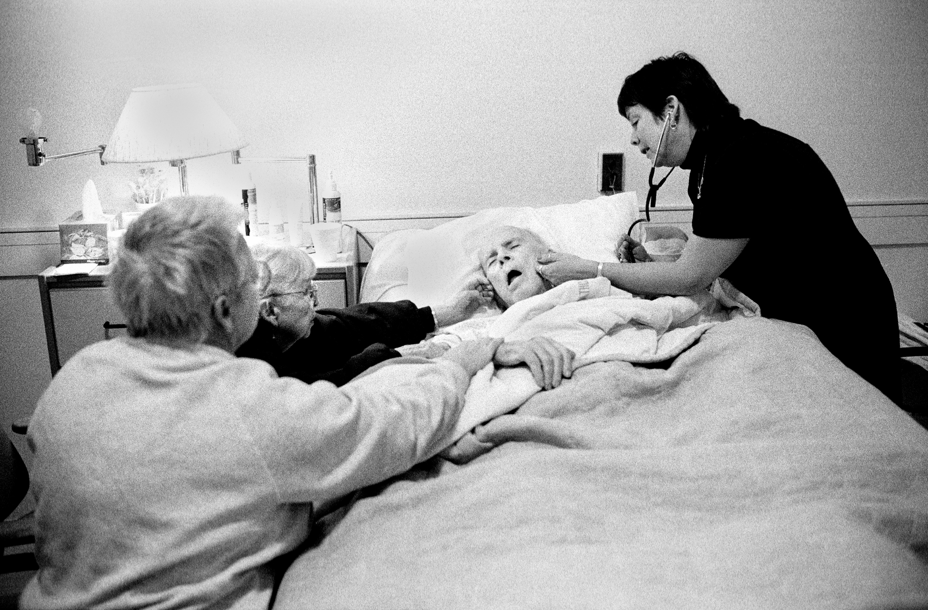 A mother and daughter sit at the side of a sick man in the hospital