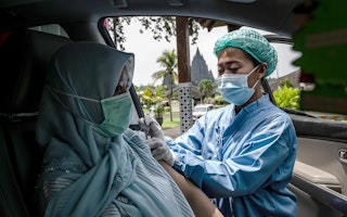 A health care worker administers a dose of a COVID-19 vaccine during a vaccination event in Yogyakarta, Indonesia, on April 5, 2021. Photo: © Ulet Ifansasti/Getty A nurse administering a vaccine to a woman sitting in a car with a temple in the background