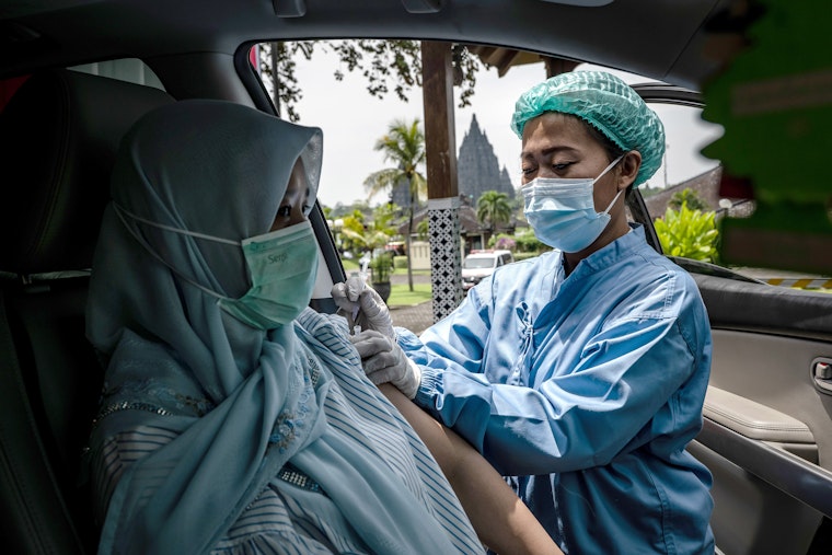A nurse administering a vaccine to a woman sitting in a car with a temple in the background