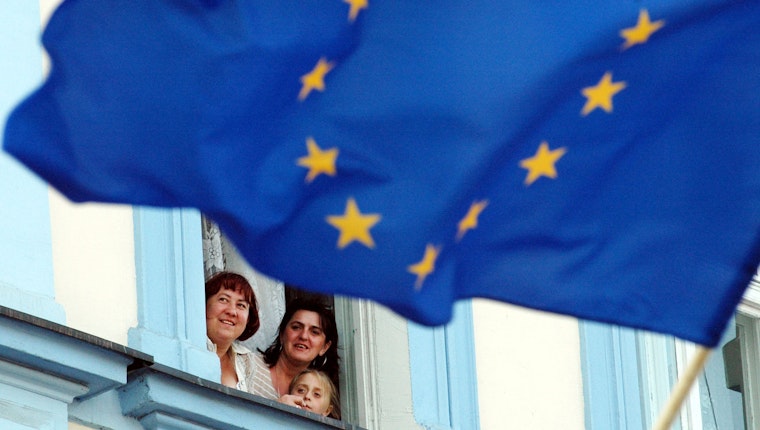 Residents look out of a window under the European flag in Slubice, Poland, on April 30, 2004. Photo credit: © Sven Kaestner/AP Photo People looking out a window behind a European Union flag