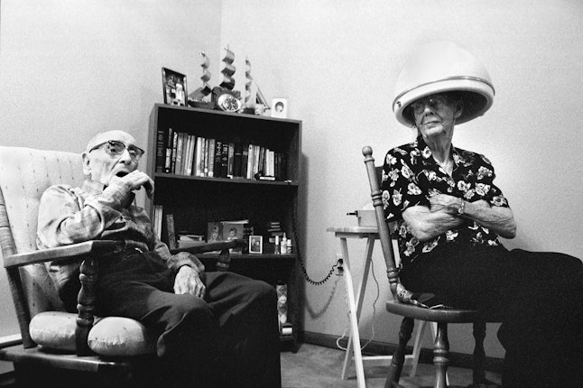Jim, 95, and his wife Lucille relax together in their room in a nursing home in Spencer, South Dakota, on August 9, 1998. © Ed Kashi/VII An older man sits on a chair next to a an older woman sitting under a hot roller machine