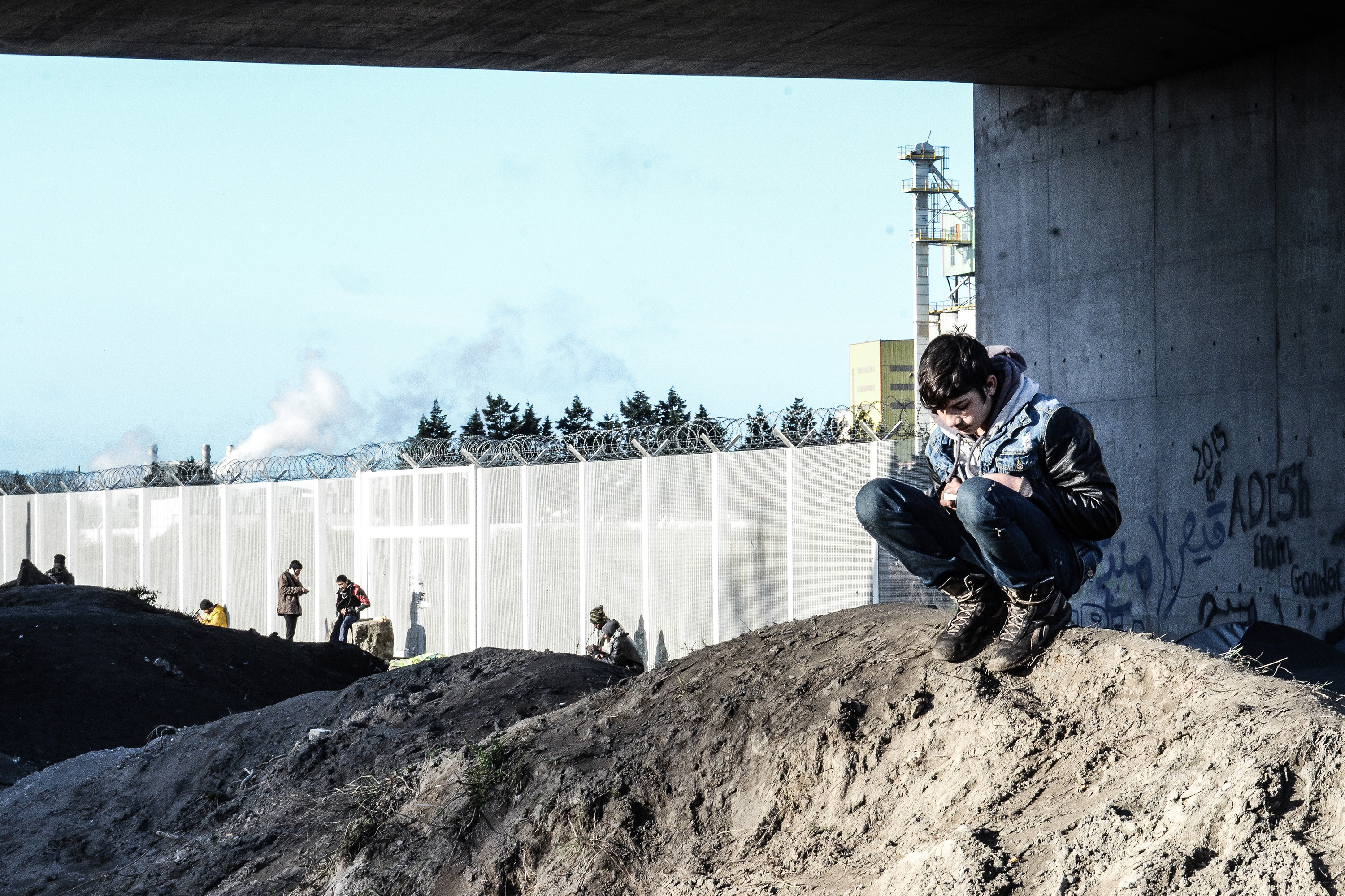 A boy squatting on a dirt pile near a tall fence