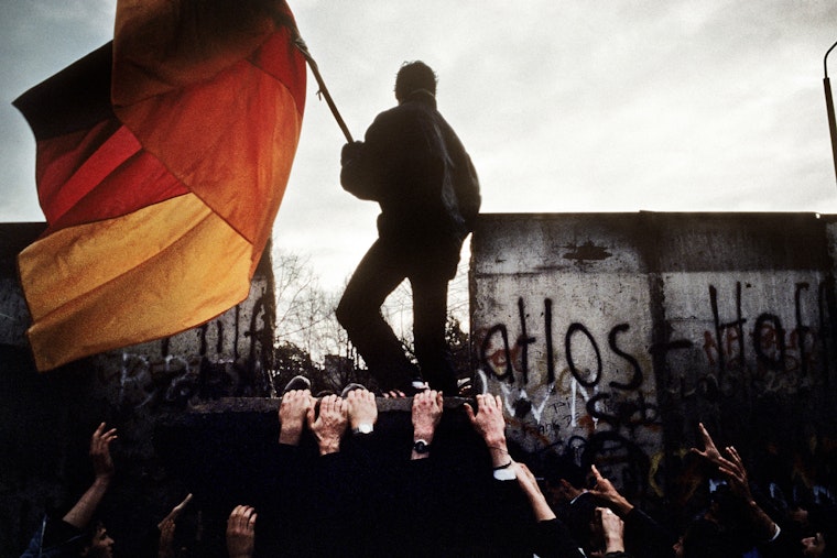 A man standing on top of a section of wall waving a large German flag.