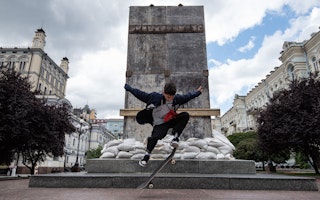 A skateboarder performs a stunt in front of a monument covered by plywood boards and sandbags