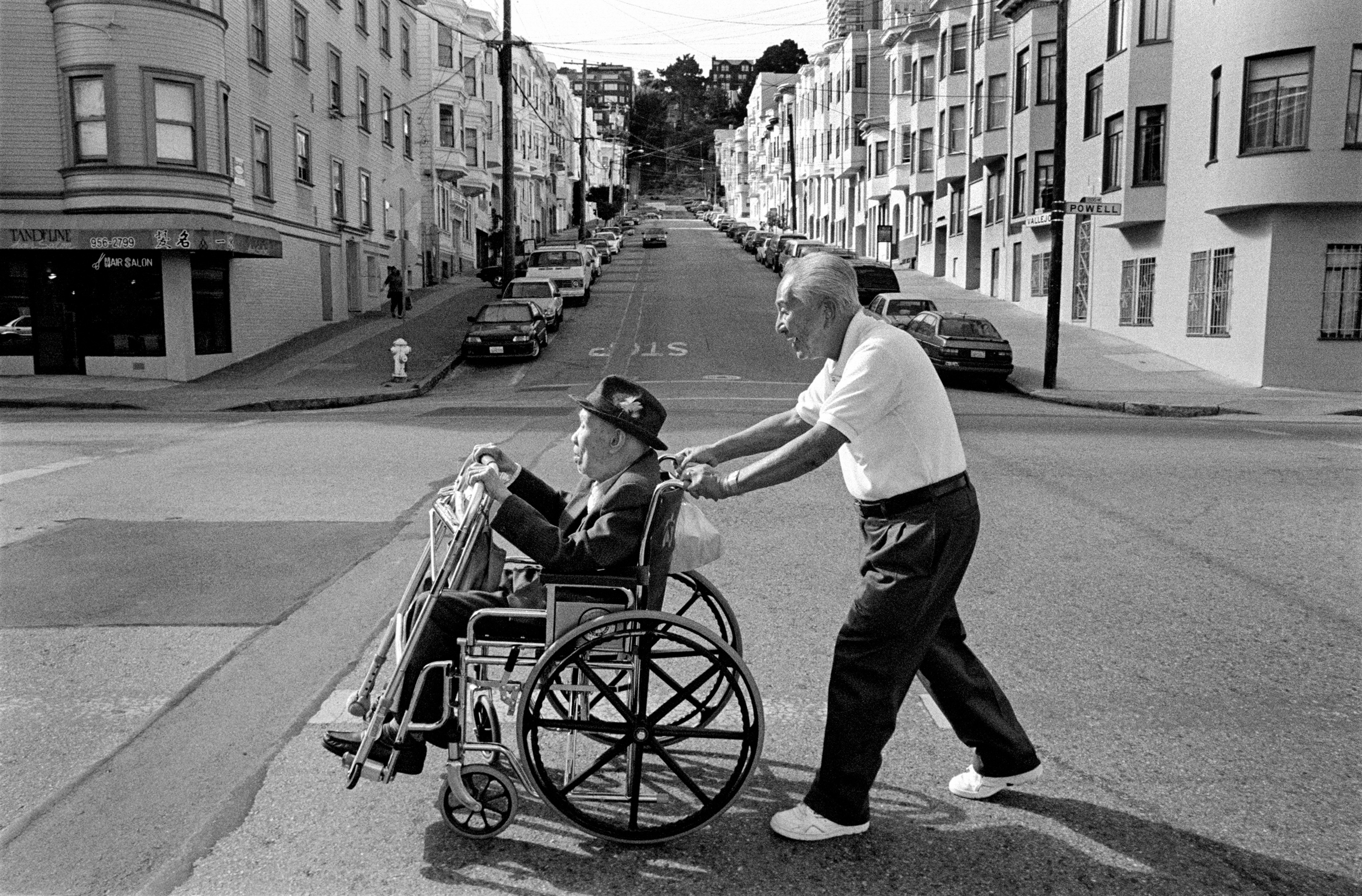 A man pushes a man in a wheelchair across the street