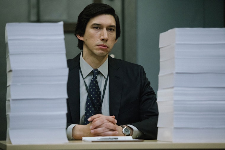 Adam Driver sitting with large stacks of paper in front of him