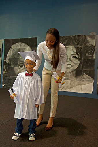Teachers stands behind small boy holding his diploma.