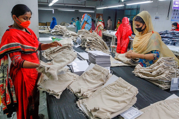 Workers fold and stack clothing at a garment factory, in Dhaka, Bangladesh, on May 7, 2020. Photo credit: © Stringer/Xinhua/Redux Two women with masks on folding clothing