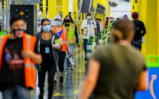 Employees return to their work stations at the Amazon Fulfillment Center in Eastvale, California, on May 20, 2020. Photo credit: © Terry Pierson/Orange County Register/Newscom People wearing vests and masks walk in a line at a warehouse