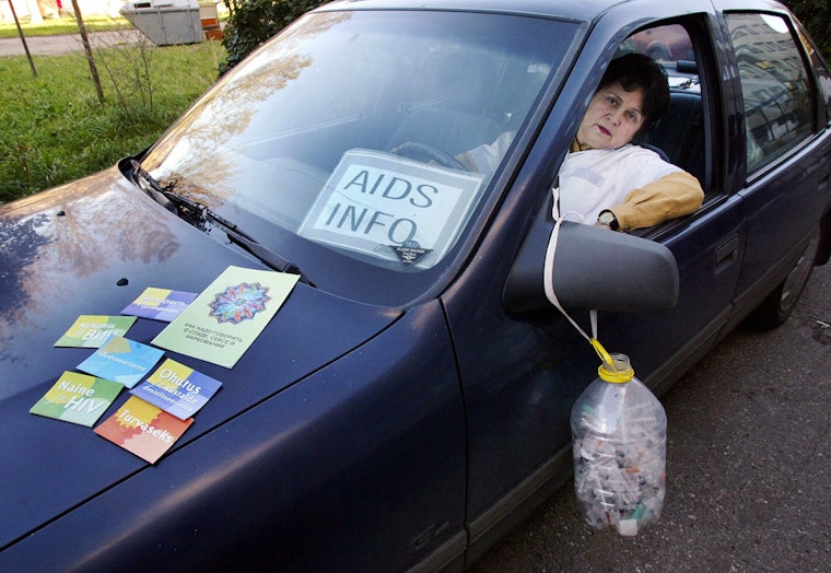 A woman sitting in the front seat of a blue car with a bucket for used needles hanging from the door