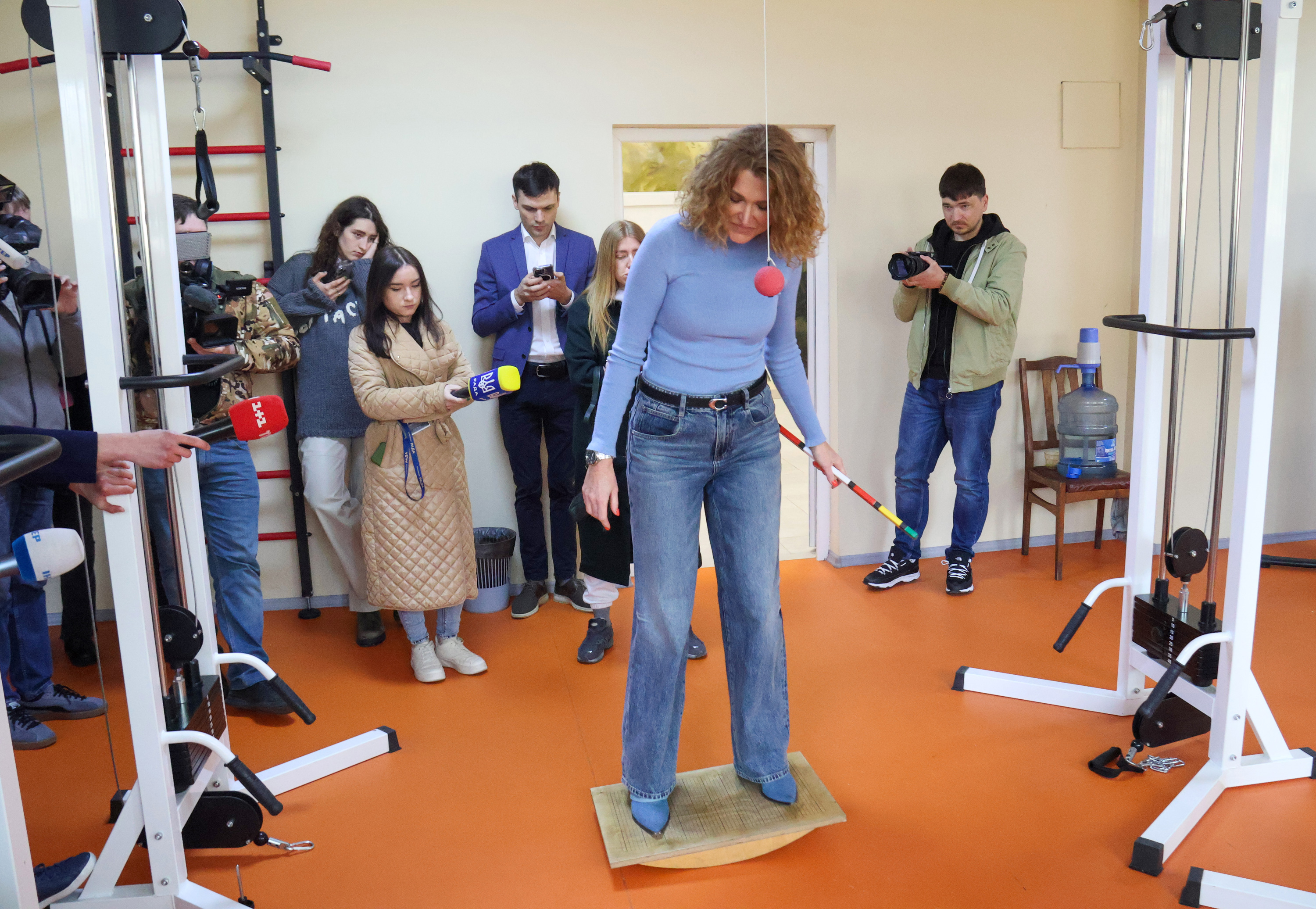 A woman stands on a balancing apparatus in the middle of a room of reporters