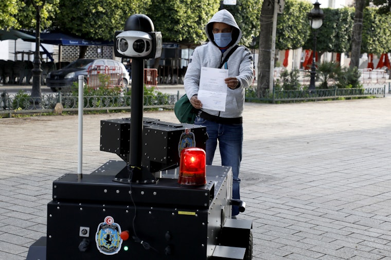 A citizen shows an exit permit to a robot that law enforcement is using to maintain an anti–COVID-19 curfew, in Tunis, Tunisia, on April 1, 2020. Photo credit: © Khaled Nasraoui/picture alliance/Getty A man holds up a piece of paper to a police robot on a public sidewalk