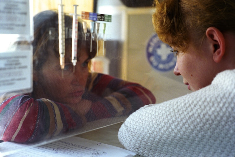 Two women lean in close to a plexiglass window where needles are displayed