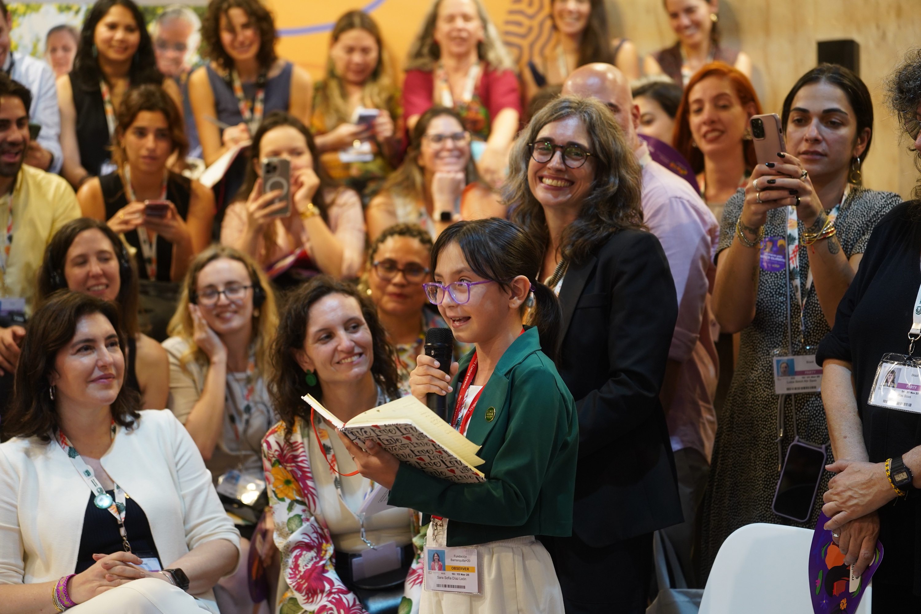 A young girl speaking into a microphone reading from a notebook in front of an audience