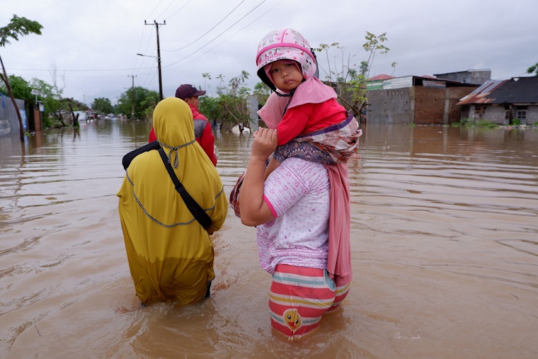 A young girl sits on the shoulders of one of three people walking through floodwaters.