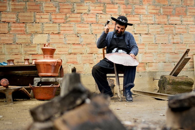 A coppersmith, who has received business support from the Roma Entrepreneurship Development Initiative, in his workshop in Sibiu, Romania, on June 15, 2021. © Cosmin I. Argeseanu for REDI A seated man holding a hammer and a piece of copper