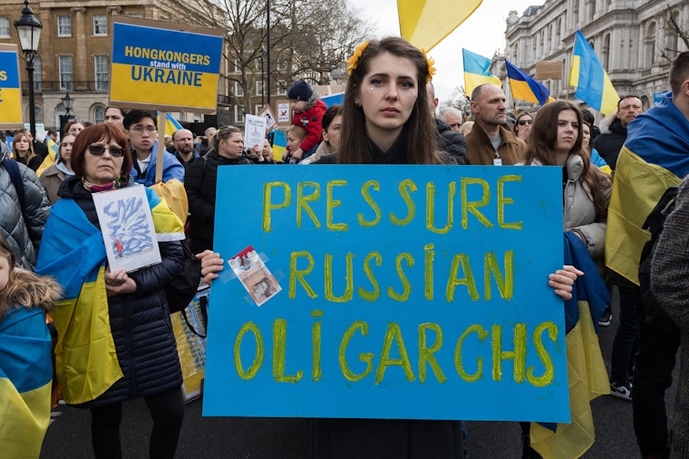 A woman at a protest holding a sign that says, "Pressure Russian Oligarchs"