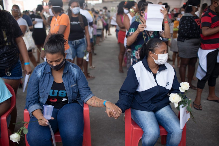 Relatives mourn for the victims of a police operation in Rio de Janeiro, Brazil, on May 13, 2021. Photo credit: © Silvia Izquierdo/AP Two people hold hands while sitting in chairs next to one another