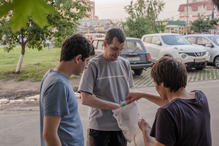 A man offers supplies to another from a bag in a parking lot