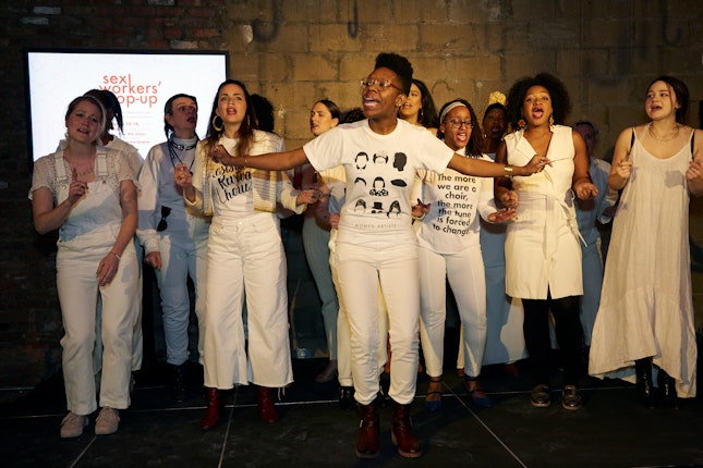 A group of women wearing white sing from a platform in an art space