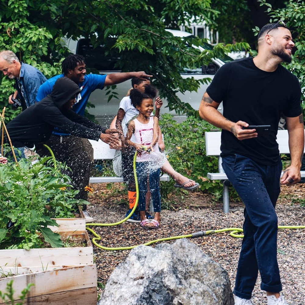 A young girl smiles and sprays a water hose in a community garden.