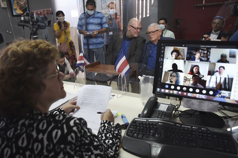 A woman sits at a desk in front of two men on a bench among a small group of onlookers and a video camera