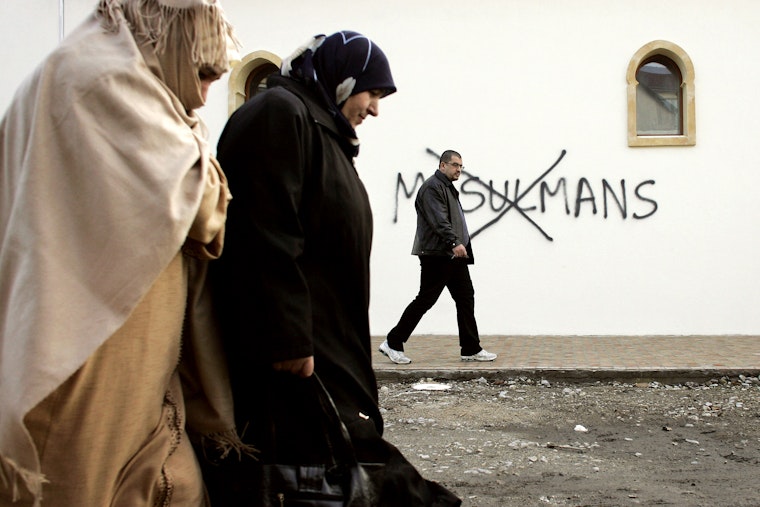 Muslim residents walk past a wall which has a racial slur painted on it in Saint-Etienne, France on February 8, 2010. Photo credit: © Laurent Cipriani/AP A group of Muslim people walk down a street, past a wall with a racial slur painted on it.