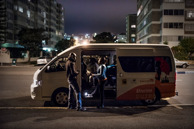 Two people stand next to a minivan which is parked under a streetlamp at night.