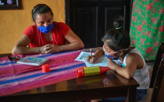 A mother watches her daughter do schoolwork at a table